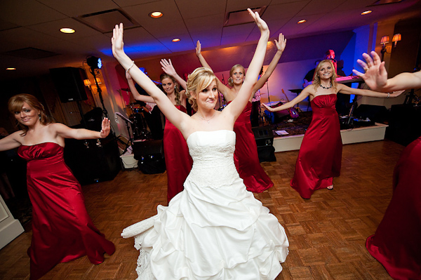 fun photo of the bride in a white ball gown dress and her bridesmaids in red full length dresses doing synchronized dancing at the reception - photo by Houston based wedding photographer Adam Nyholt 